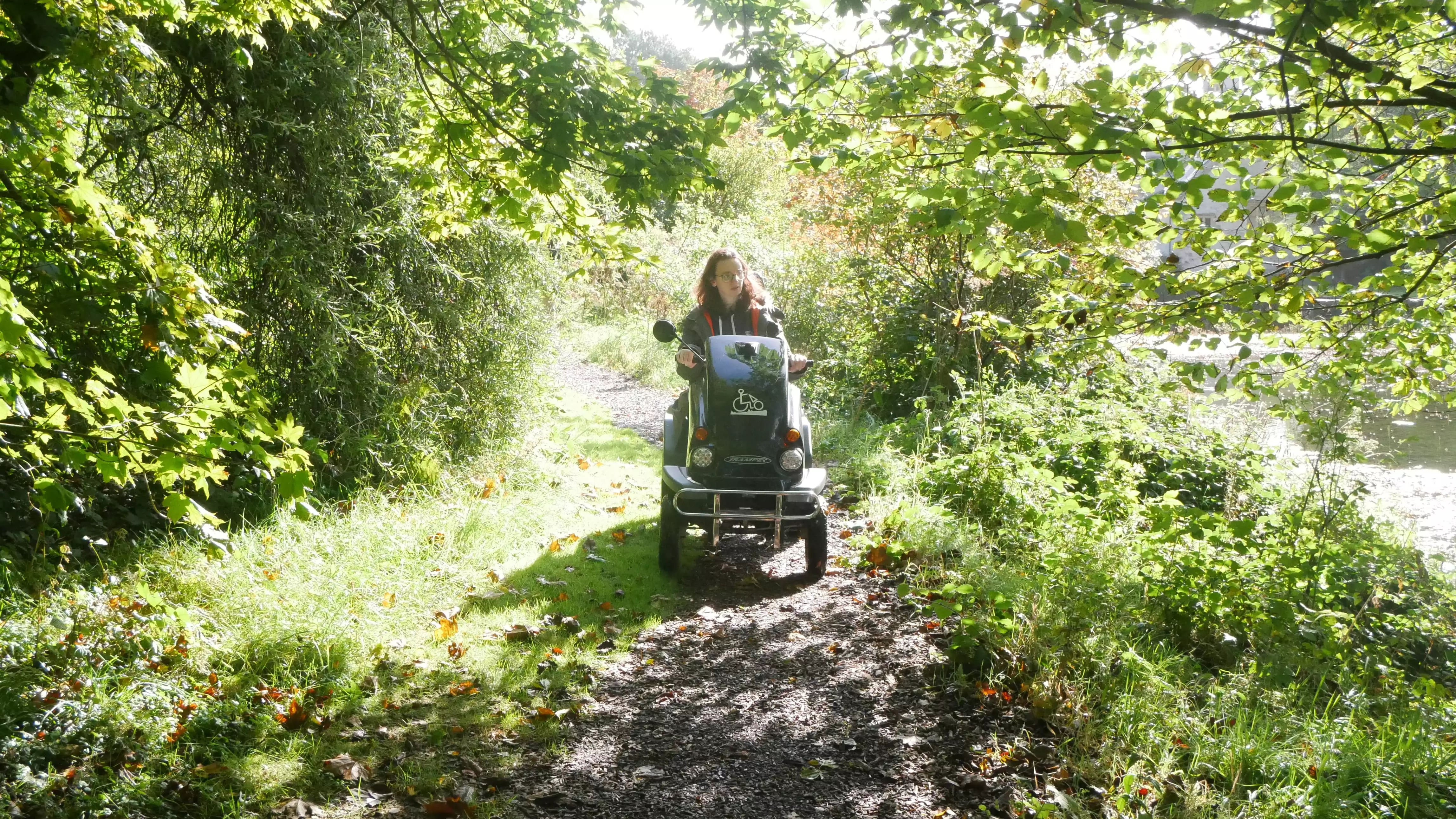 Woman on a mobility scooter built for muddy paths in a wooded leafy area.