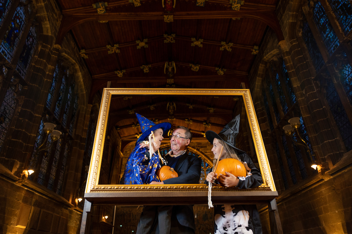 In a dark great hall within a frame there are two little girls dressed as witches and holding pumpkins with an older man in the middle in between the girls.