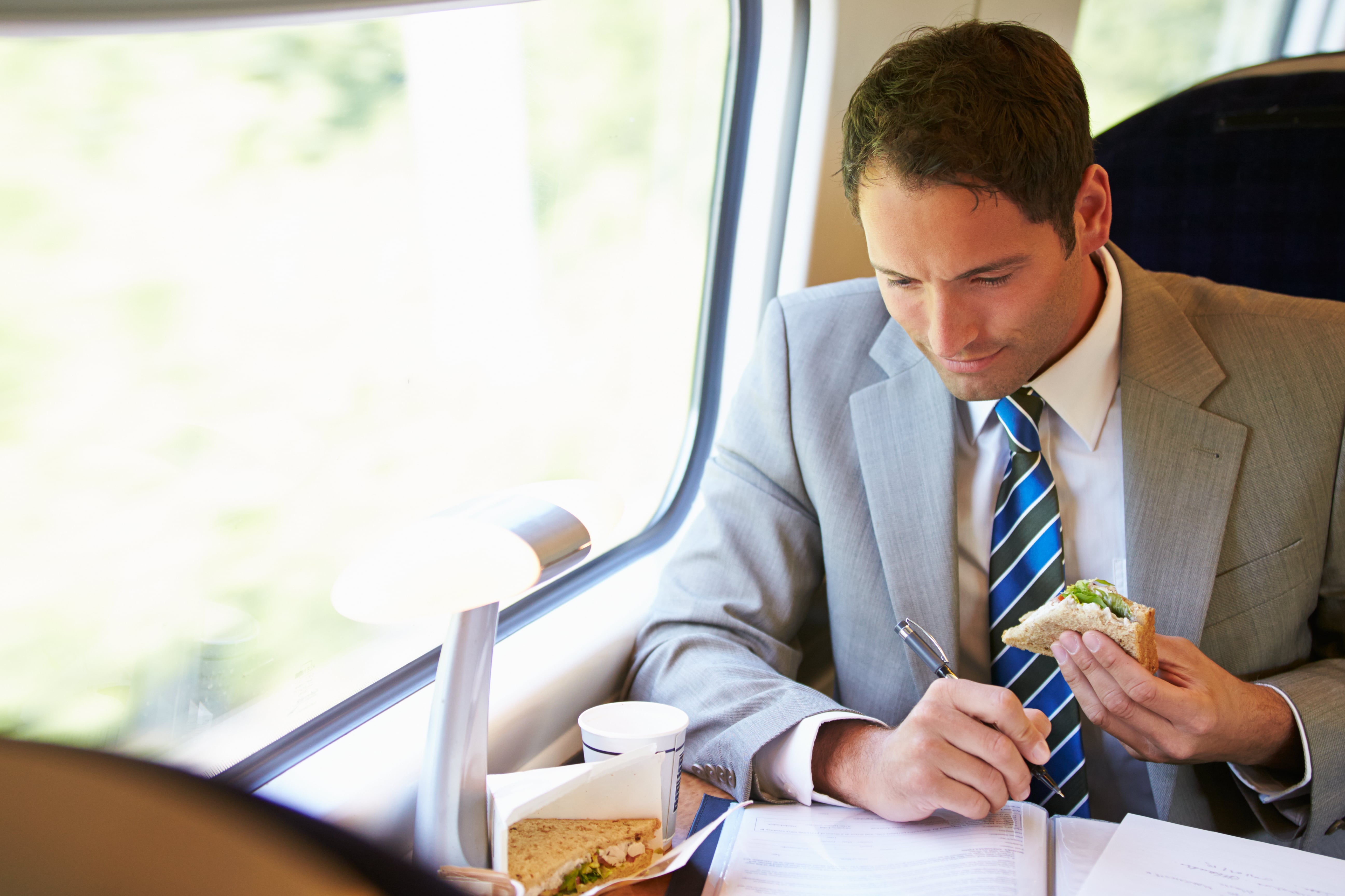 Food on Trains: A young businessman enjoys a sandwich while completing paperwork as he sits at a train window seat