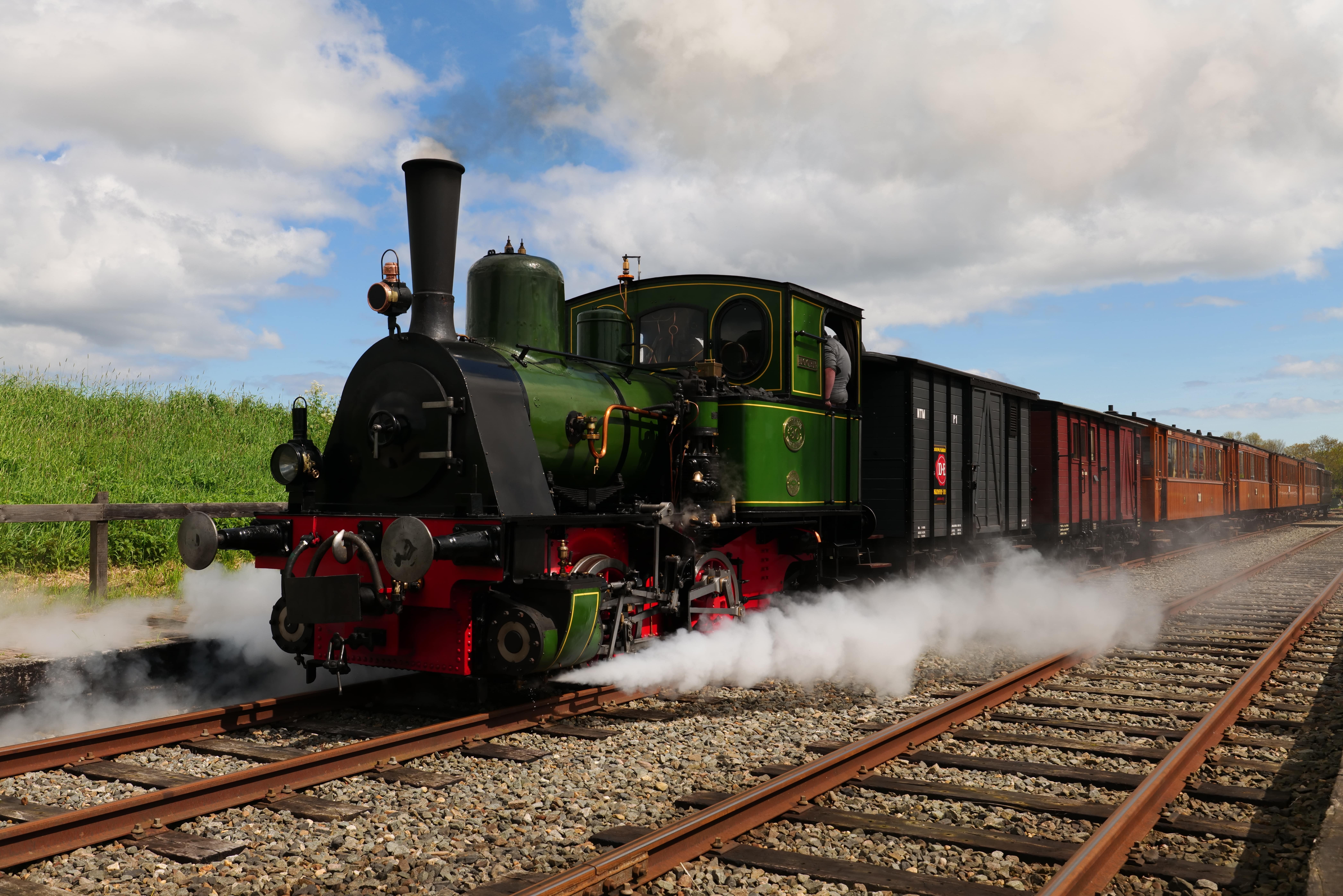 Rail Advertising: A vintage green steam locomotive with black and red detailing pulling classic wooden passenger carriages, releasing steam as it travels down the railway