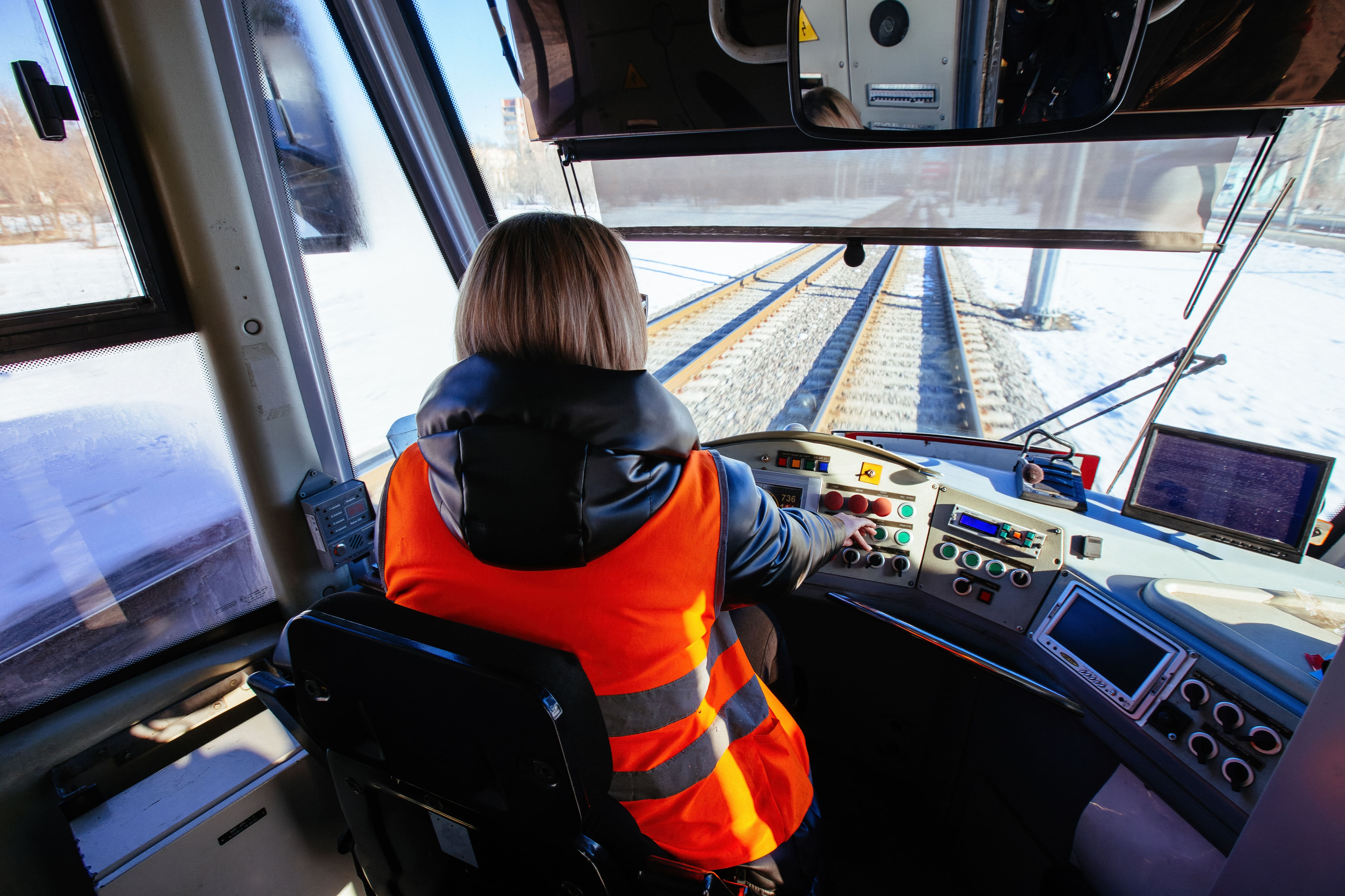 West Coast Train Line: The back of a woman’s head as she wears a hi-vis vest while driving a train forward.