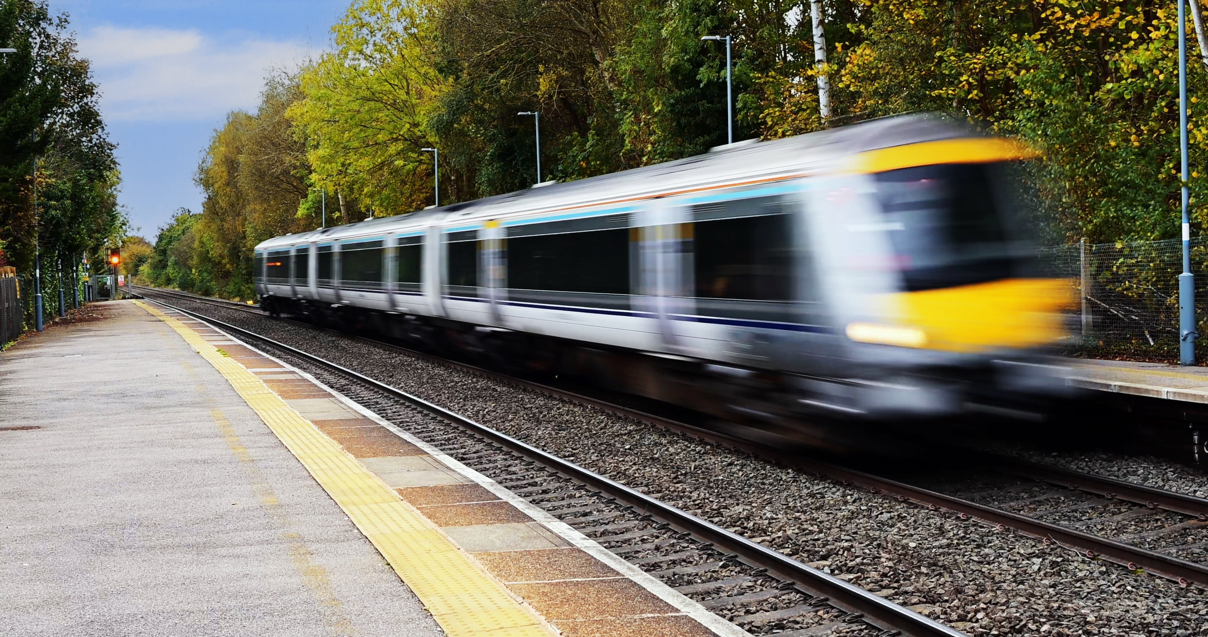 West Coast Train Line: A train speeds past an empty platform, with trees on one side of the station.
