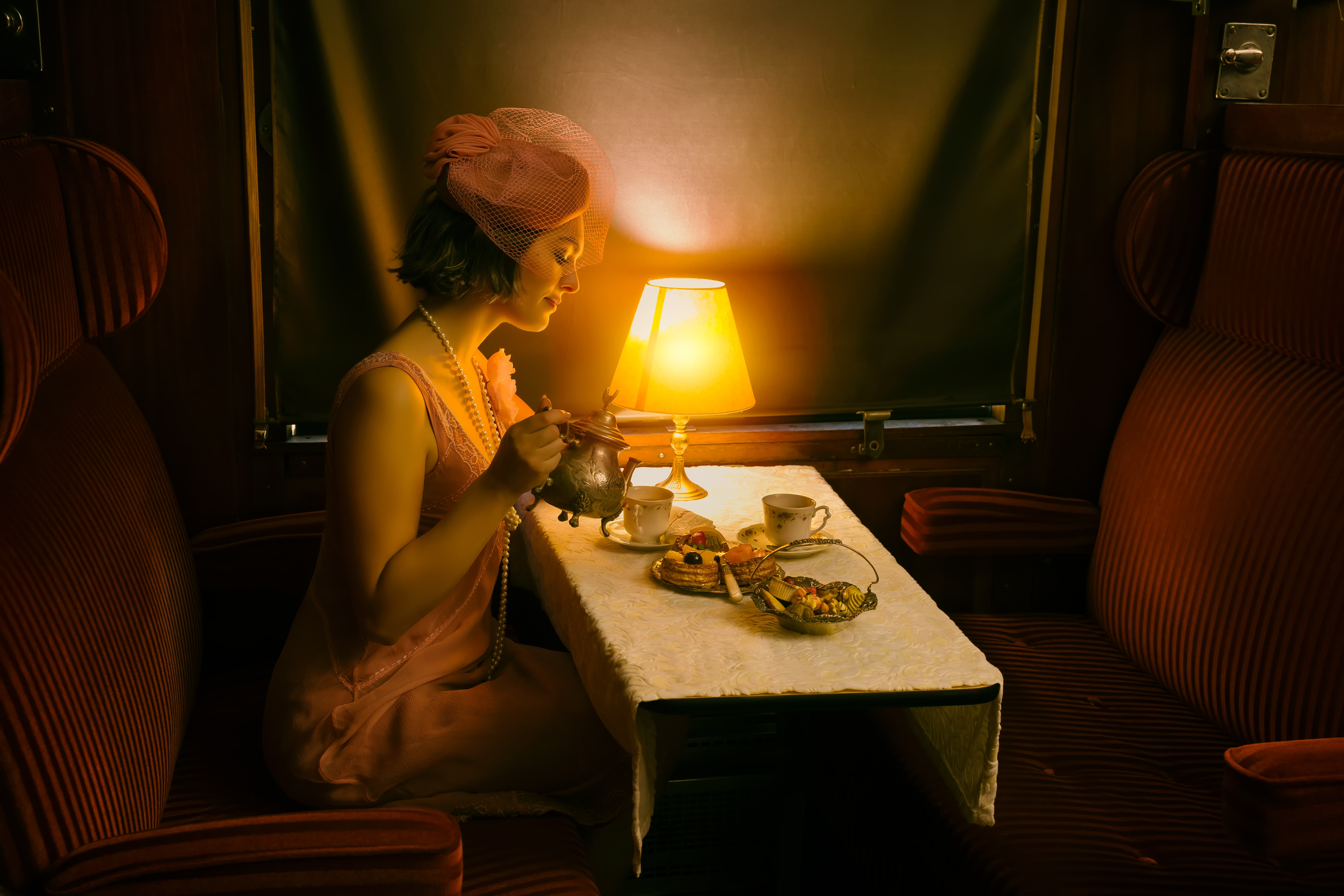 Rail Queens: An elegant woman in vintage attire enjoying tea and pastries in a dimly lit retro train compartment, illuminated by a warm table lamp