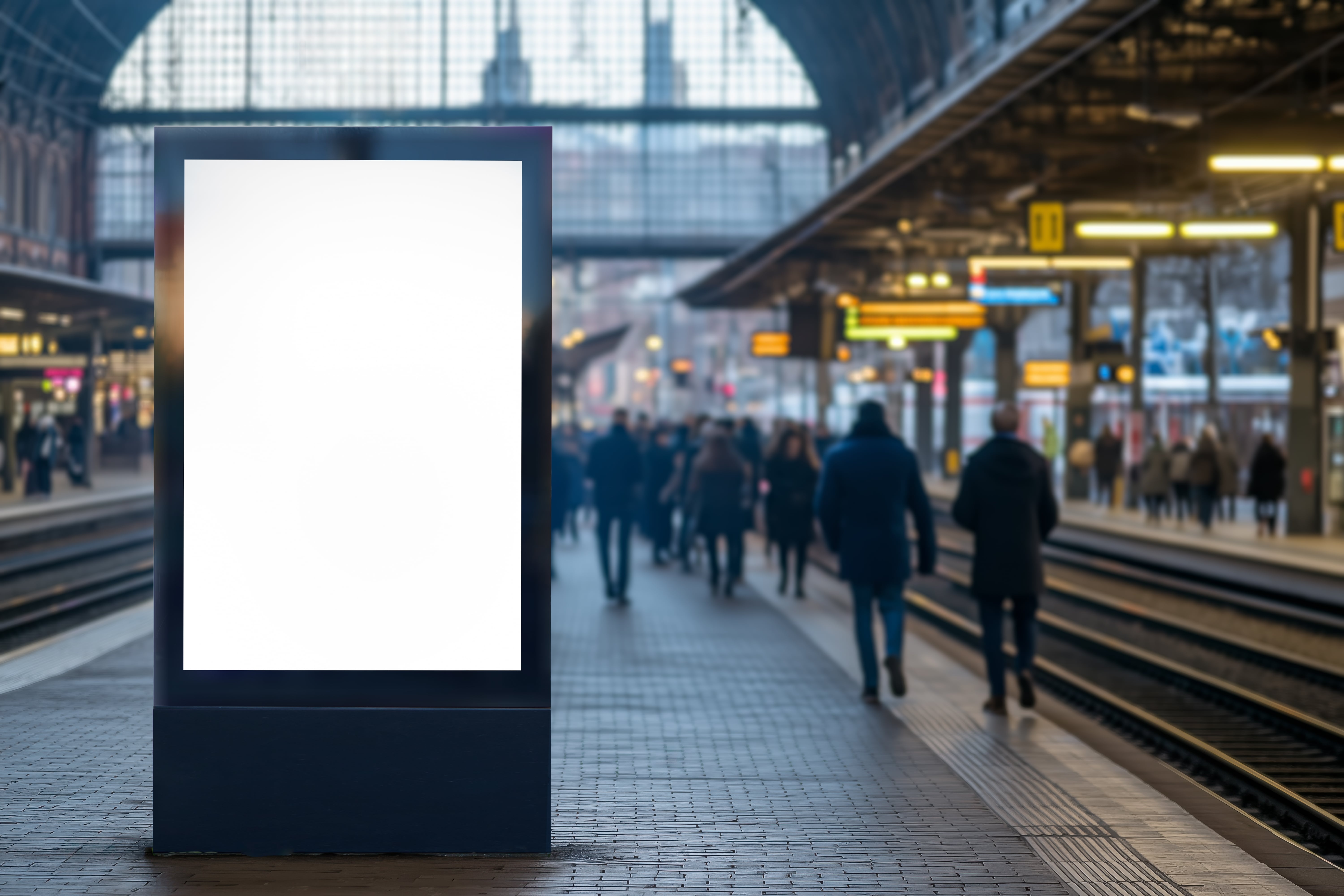 Trains in the UK: A blank digital billboard at a train station with many passengers walking by in the distance.