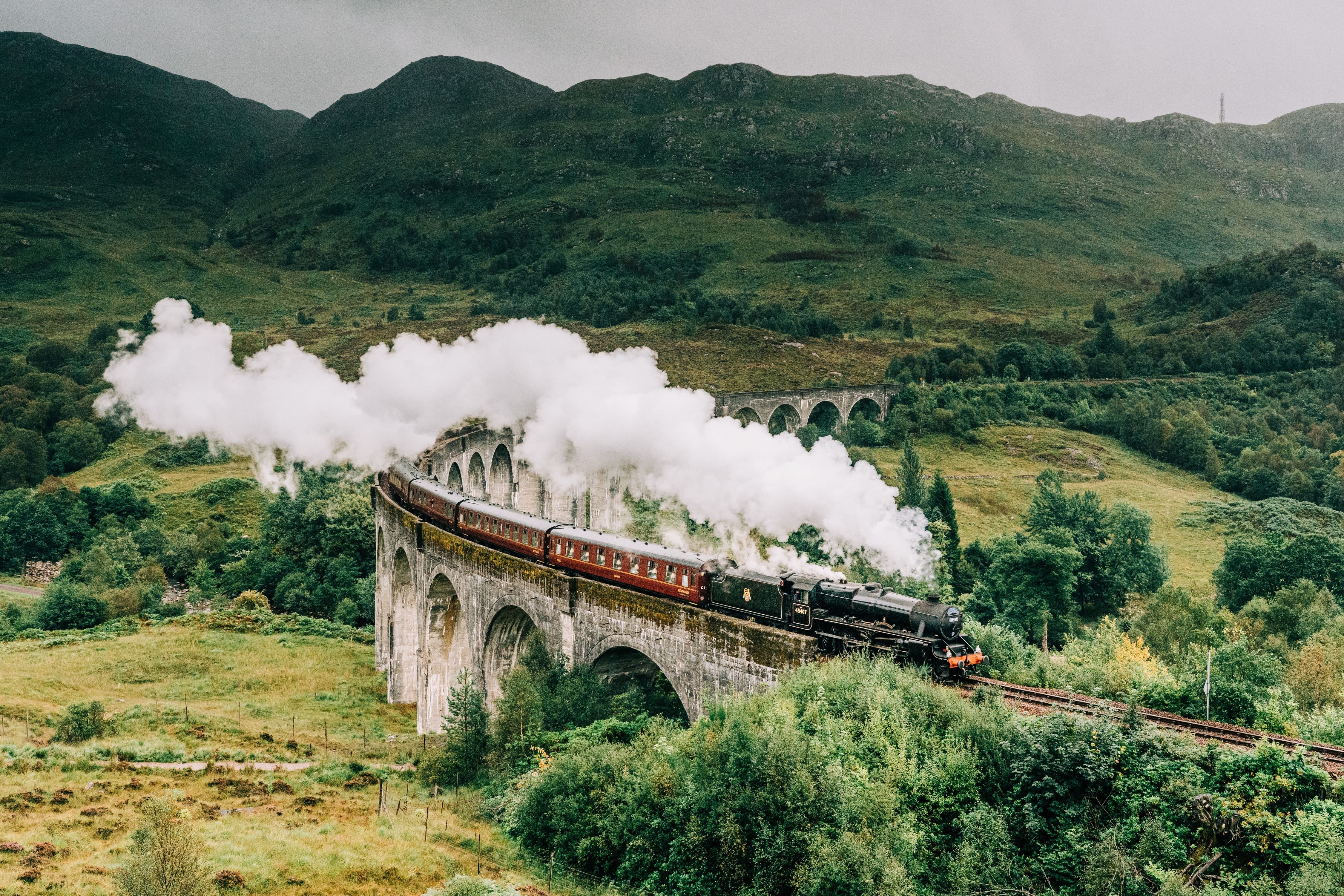 Trains in the UK: A steam train rides through a lush valley over an aqueduct as steam emerges from its chimney.