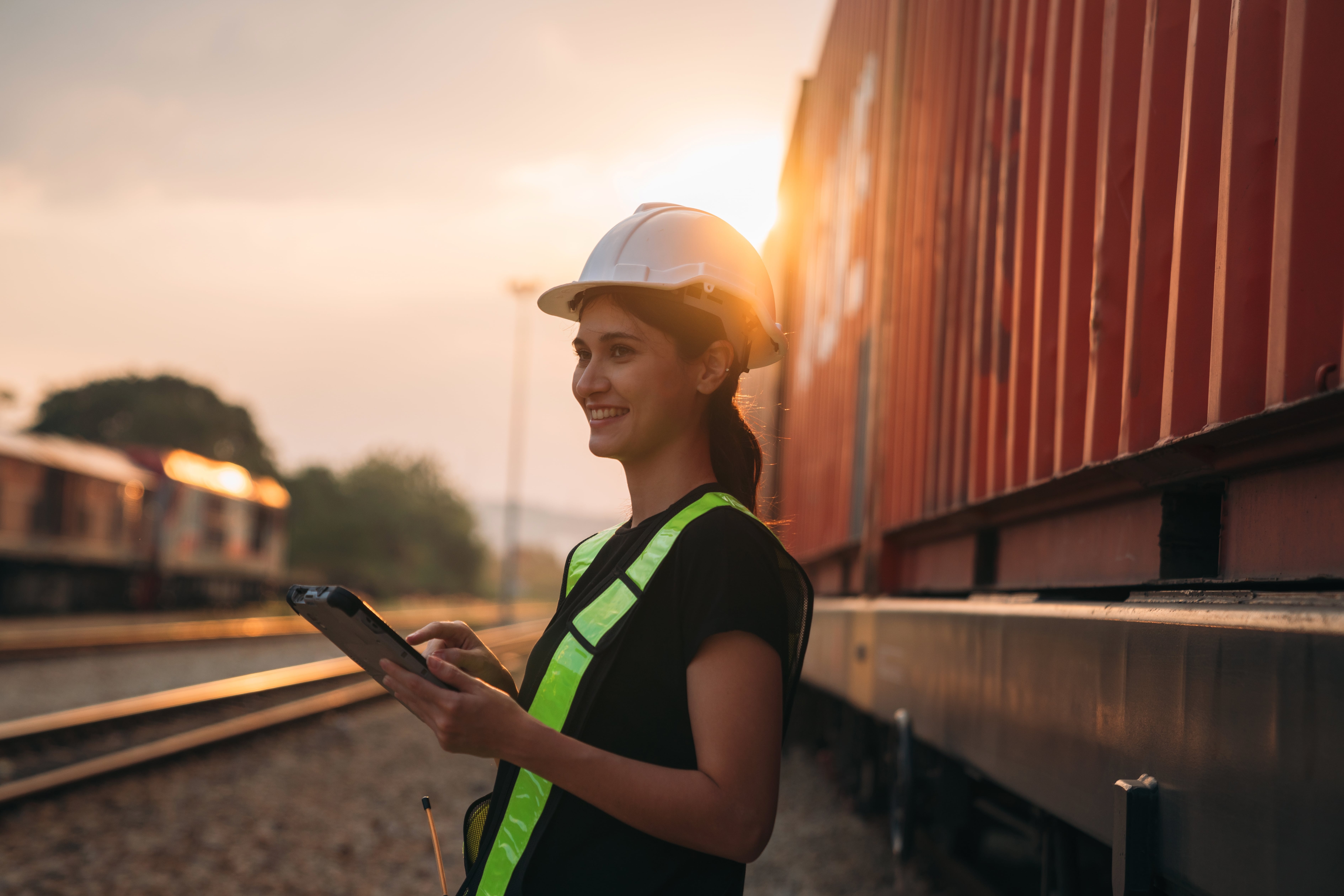 Jobs in Rail: Smiling female railway worker wearing a hard hat and high-visibility vest, holding a tablet while inspecting a freight train at sunset