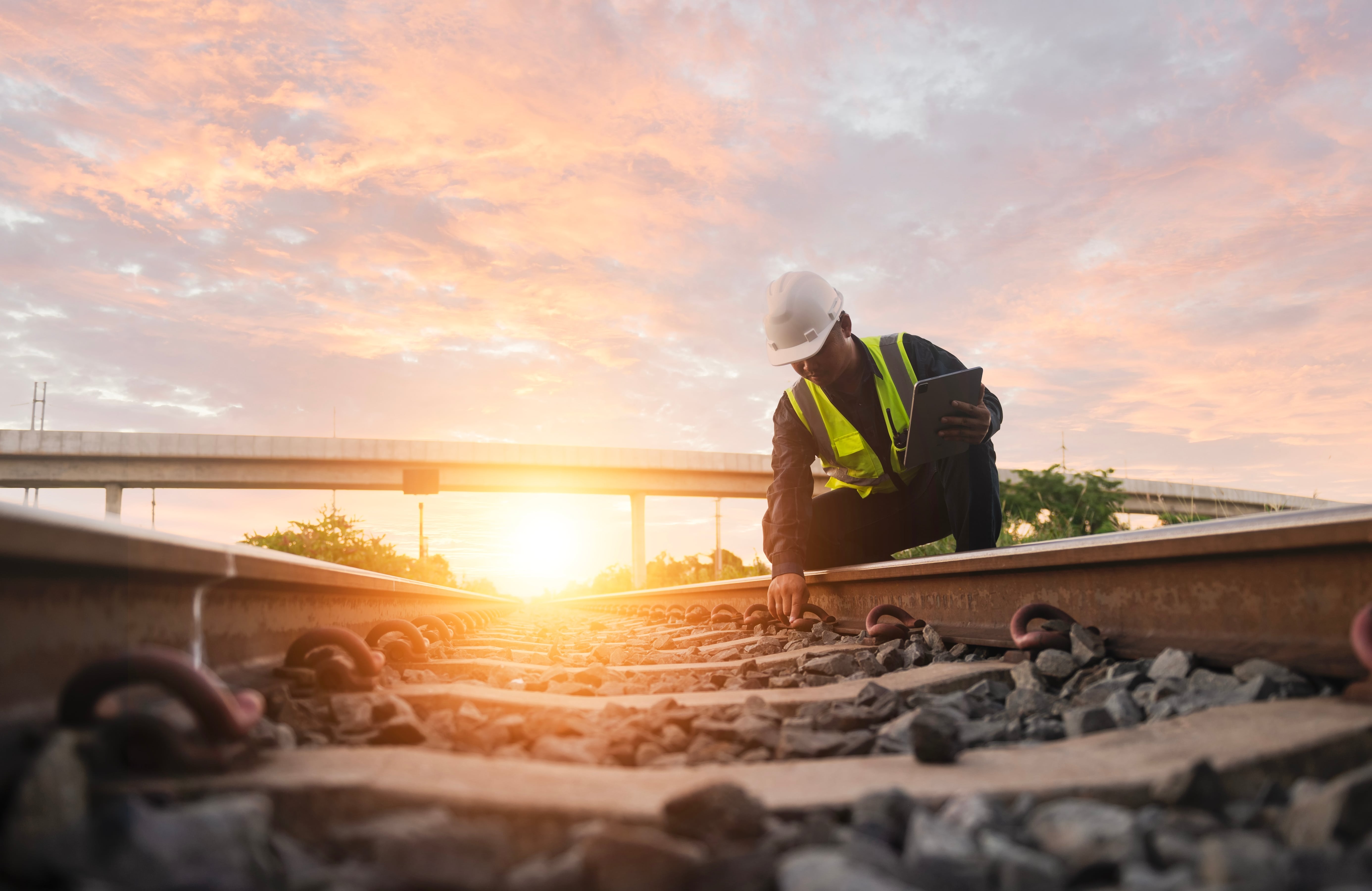 Jobs in Rail: Railway worker in a hard hat and high-visibility vest inspecting train tracks at sunset.
