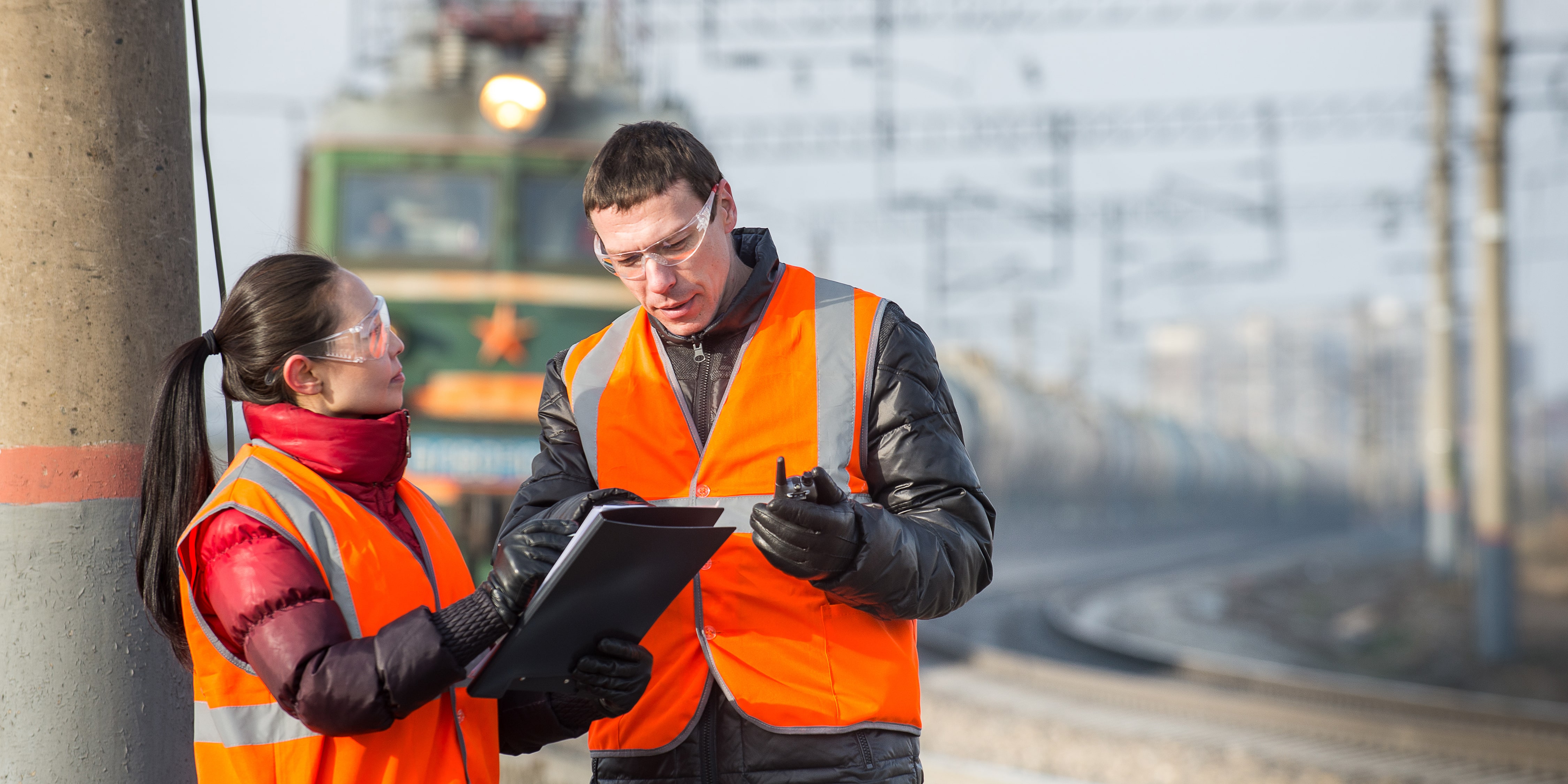 Jobs in Rail: Two railway workers in high-visibility vests and safety goggles discussing a clipboard report near train tracks, with a locomotive approaching in the background