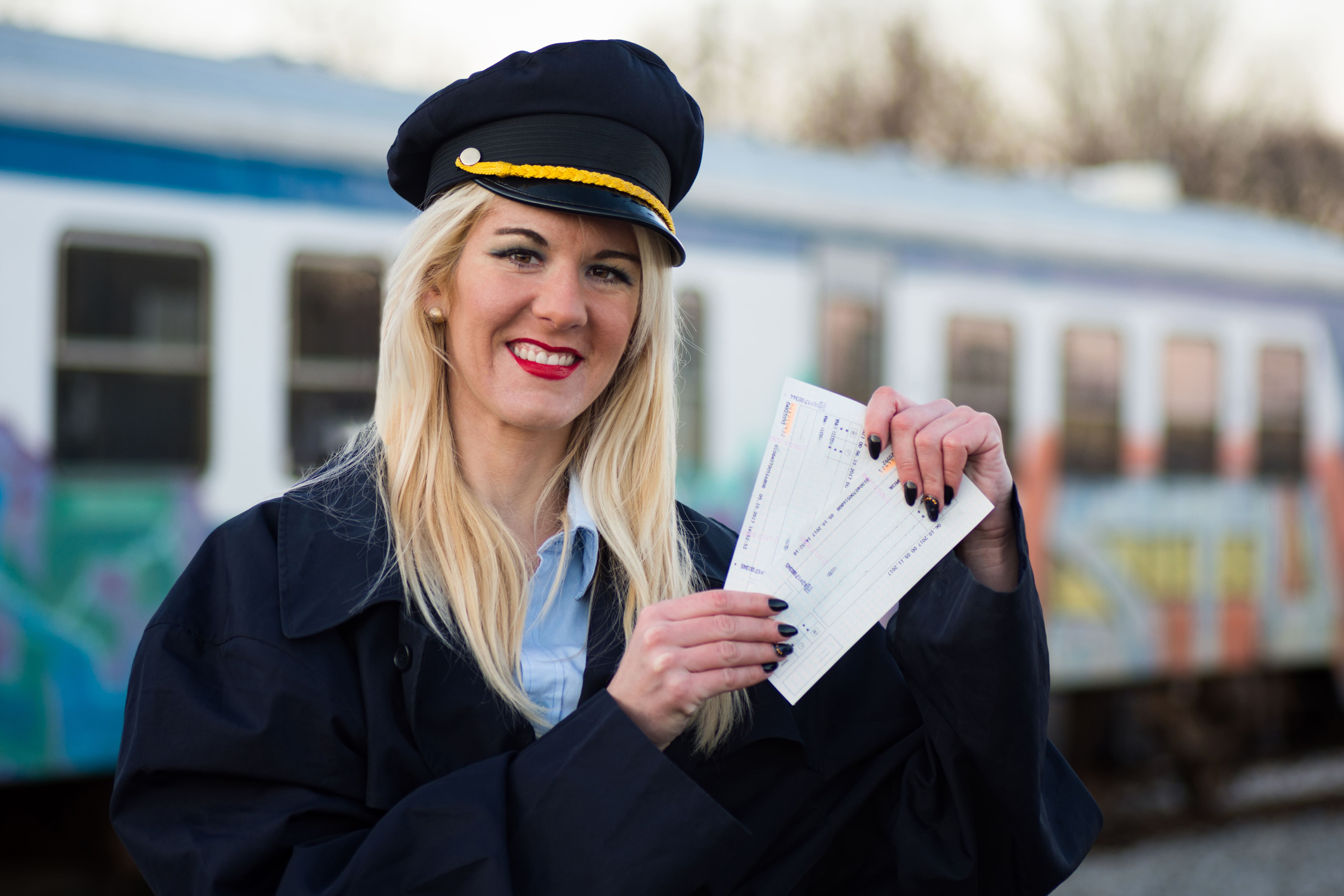 Women in Rail: A blonde-haired woman wearing a train conductor’s hat holds up two tickets, as a train remains stationary in the background