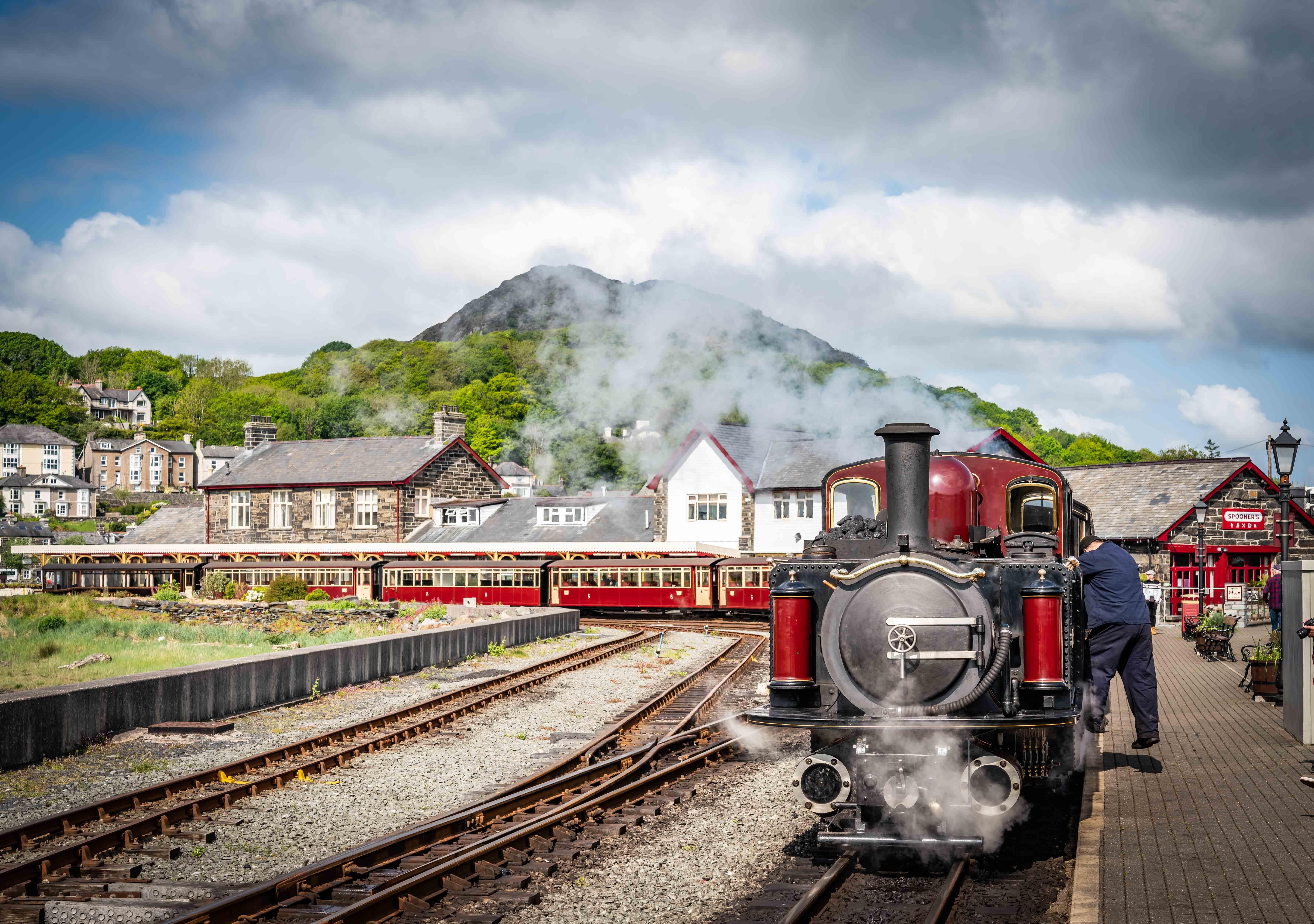 Bicentennial: A steam locomotive at the Ffestiniog and Welsh Highland Railways station, with smoke rising against a scenic backdrop of mountains and historic buildings