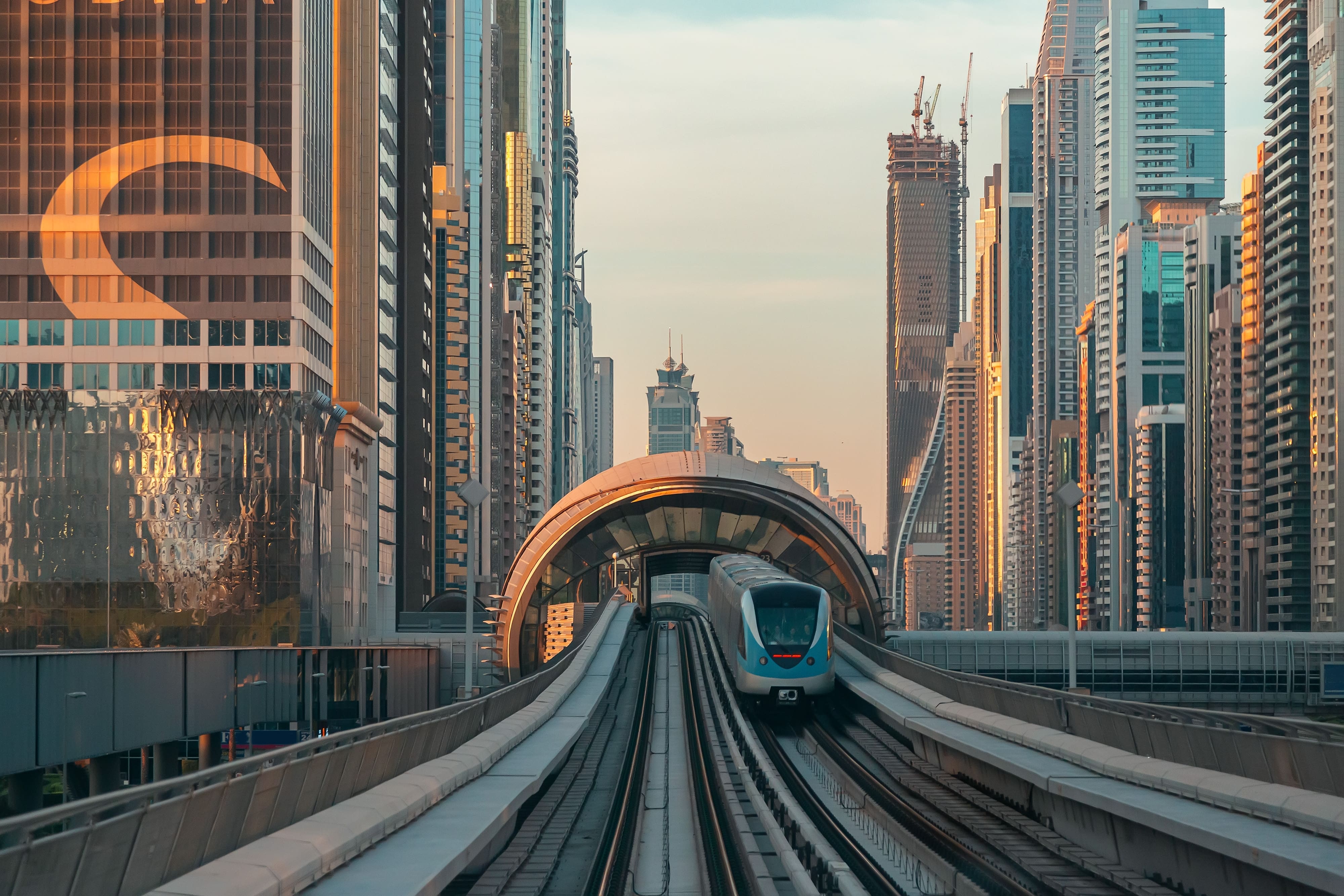 Future Trains: In the distance, a train comes towards the camera on an overground railway line, as the sun sets on a city in the background. Future Trains: An engineer peers under a stationary train as they contemplate it.