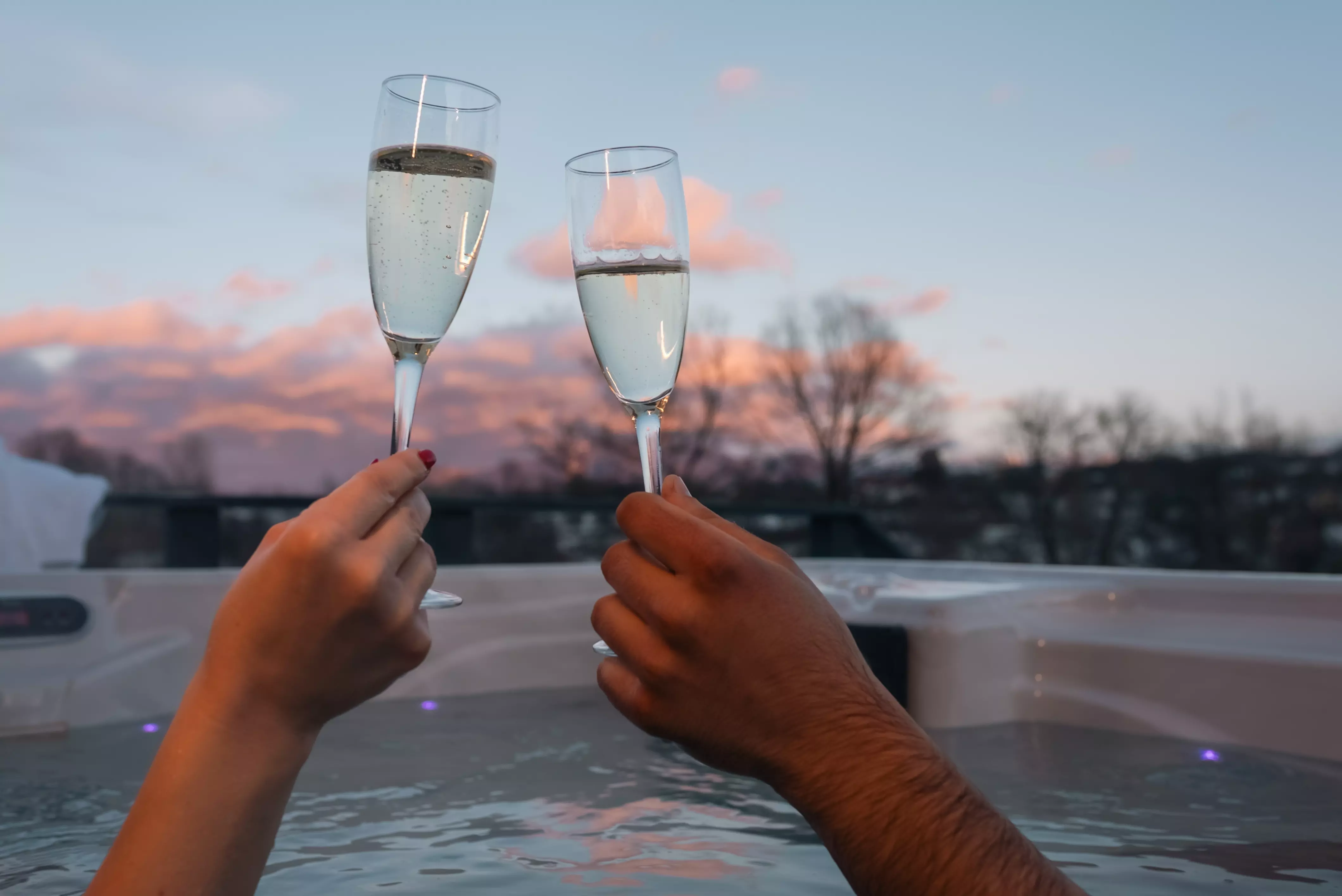 Closeup of hands clinking with champagne in outdoor hot tub at sunset.