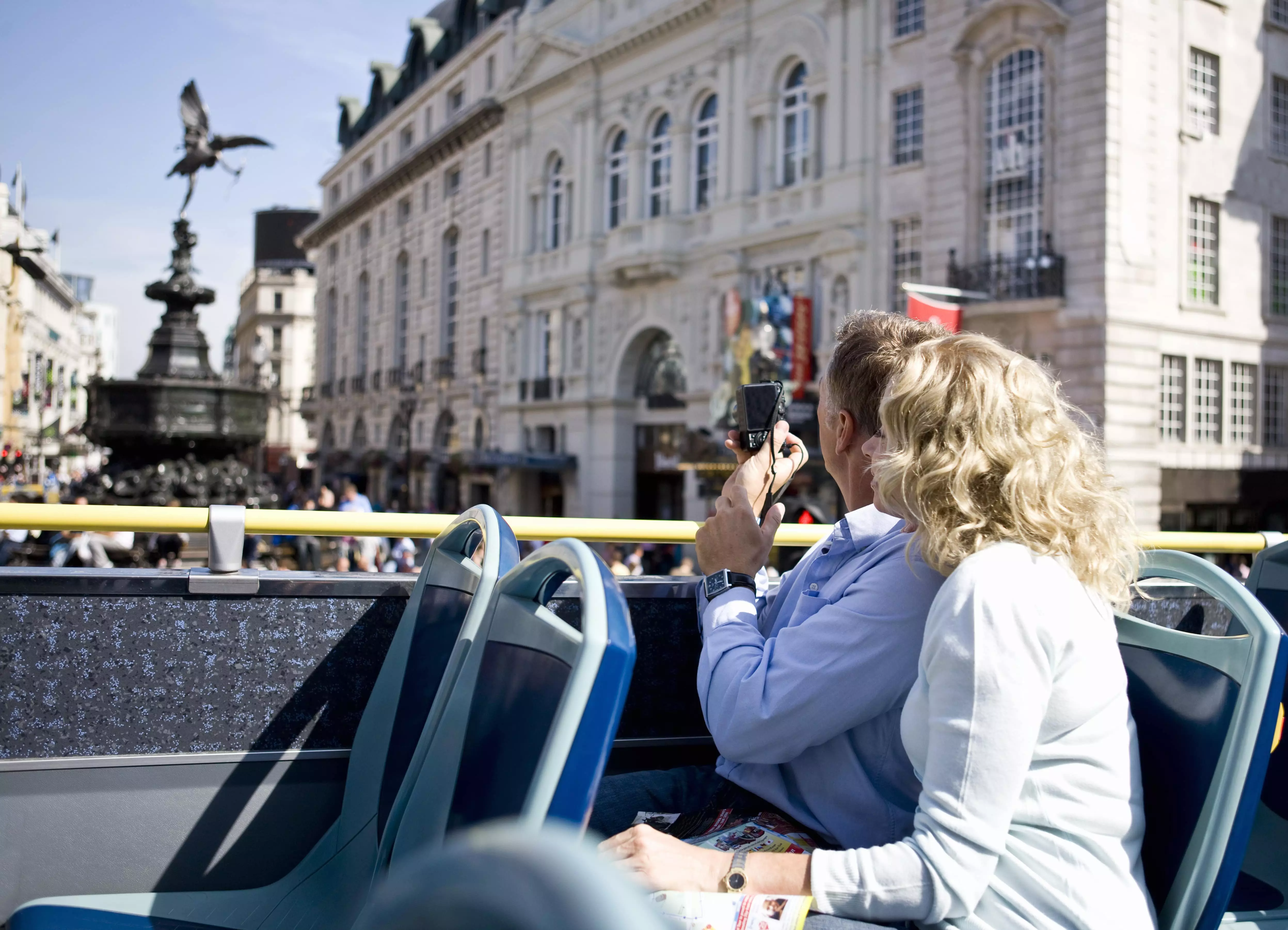 A middle-aged couple sitting on a London sightseeing bus, taking photographs of Piccadilly Circus